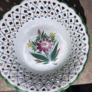 May include: White ceramic decorative plate with a green scalloped edge and openwork design. The plate features a floral motif with a large purple flower, green leaves, and accents of blue and yellow. The plate is approximately 10 inches in diameter.