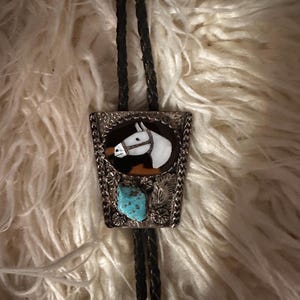 May include: A bolo tie featuring a silver-toned metal slide with a central inlay of a white horse head silhouette against a dark background. Below the horse, a rough-cut turquoise stone is set alongside silver leaf and floral accents.