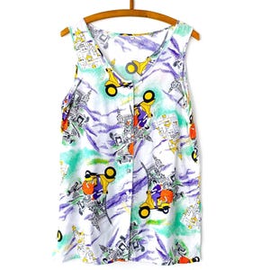 May include: A sleeveless white top with a colorful print of yellow scooters, buildings, and abstract shapes. The top has a V-neck and is hanging on a wooden hanger.