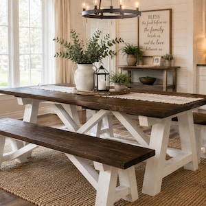 May include: A rectangular dining table with a dark brown wooden top and white X-frame legs, accompanied by two matching benches. A white pitcher with greenery, a black metal lantern, and a white table runner are placed on the table surface.