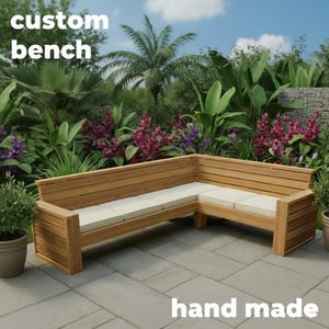 Peut inclure: Banc en bois en forme de L avec coussins beiges. Le banc est en bois brun clair et comporte des lattes horizontales sur le dossier. Les mots "custom bench" et "hand made" sont visibles. Le banc est à l'extérieur, entouré de plantes.