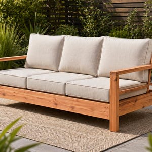 Puede incluir: Un sofá de exterior de tres plazas con estructura de madera marrón claro y cojines beige. El sofá tiene forma rectangular con respaldo bajo y reposabrazos. Los cojines tienen un ribete gris claro. El sofá está colocado sobre una alfombra tejida.