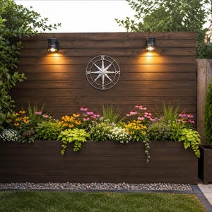 May include: A dark brown horizontal wooden slat wall featuring a central silver metal compass rose wall art piece, two illuminated wall sconces, and a large rectangular planter box filled with colorful yellow, pink, and white flowers.