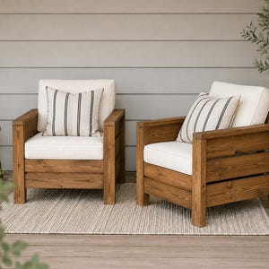 May include: Two armchairs constructed from dark stained wood with thick, off-white seat and back cushions. Each chair features a square frame design and includes a decorative throw pillow with vertical black stripes on an off-white background.