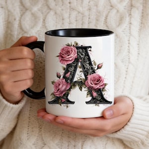 May include: White ceramic mug with a black interior and handle, featuring a floral design with the letter "A". The letter is black with ornate detailing, surrounded by pink roses and green leaves. The mug is held by a person wearing a cream-colored sweater.