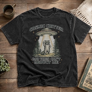 May include: A charcoal gray mineral-washed t-shirt featuring a graphic of a raccoon making a peace sign under a UFO beam. The text reads COSMIC CRITTER and BAD VIBES CAN'T ABDUCT ME. The design includes stylized pine trees and small stars.