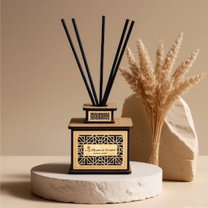 Puede incluir: Un difusor de aceites esenciales de madera con varillas negras. El difusor tiene un diseño geométrico y el texto "Maison de Fraicheur NATURAL SCENT". El difusor está sobre un pedestal blanco con trigo seco y una piedra en el fondo.