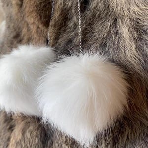 May include: White fluffy pom-pom earrings suspended from silver chains. The earrings are round and have a soft, textured appearance. The background is a mix of brown and grey fur.