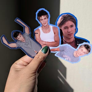 May include: Four die-cut stickers featuring images of a young man in various poses. The stickers have a blue border and are held by a hand with green glitter nail polish. The man is wearing a gray shirt, a white tank top, and a gray t-shirt.