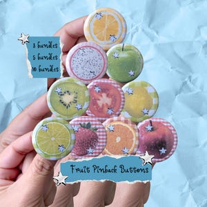 Könnte beinhalten: Eine Sammlung runder Ansteckbuttons mit verschiedenen Fruchtmotiven wie Zitrone, Drachenfrucht, grünem Apfel, Kiwi, Tomate, Erdbeere, Orange und Apfel. Jeder Button ist mit kleinen silbernen Sternen verziert. Der Text lautet 3 bundles, 5 bundles, 10 bundles und Fruit Pinback Buttons.