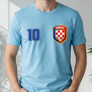May include: Light blue t-shirt with the number 10 in blue on the left side. A Croatia emblem is on the right, featuring a red and white checkered shield, a gold soccer ball, and the word "CROATIA".