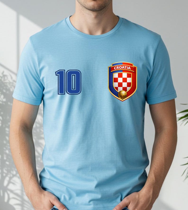 May include: Light blue t-shirt with the number 10 in blue on the left side. A Croatia emblem is on the right, featuring a red and white checkered shield, a gold soccer ball, and the word "CROATIA".