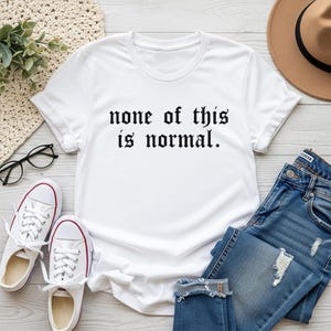 May include: A white t-shirt with the phrase "none of this is normal" in black gothic font. The shirt is styled with blue denim jeans, white trainers with red trim, and a brown hat. Black glasses and a woven mat are also visible.
