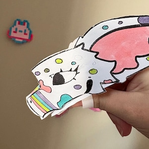 May include: A handmade paper cutout of a cartoon dragon head, featuring a white base with pink, blue, and yellow accents. The dragon's eye is black, and a rainbow design is visible on the lower edge. A pixelated character is in the background.
