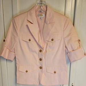 Pode incluir: Um blazer rosa claro com mangas curtas, quatro bolsos frontais e botões dourados. O blazer está pendurado em um cabide transparente. As mangas estão dobradas. A etiqueta da marca é visível.