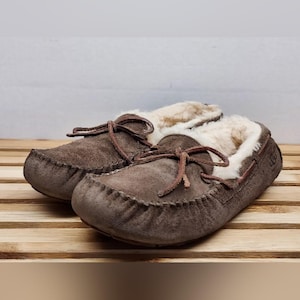 May include: A pair of brown suede moccasin slippers with a soft, cream-colored interior lining. The slippers have a leather tie detail and are displayed on a wooden surface. The UGG logo is visible on the side of one slipper.