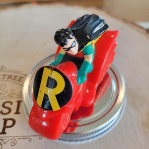 May include: A red toy motorcycle with a black and yellow "R" logo on the front. A small Robin figure with a black cape and green gloves is riding the motorcycle. The toy is on a silver lid.