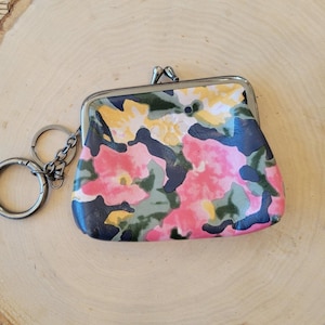 May include: A small, floral-patterned coin purse with a metal clasp and attached keychain. The purse features a colorful design with pink, yellow, green, and navy blue floral patterns. The purse is set against a light brown wooden background.