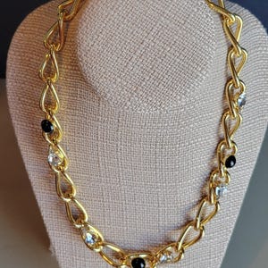 May include: A gold-coloured chain necklace with a linked design. The necklace features alternating clear and black gemstone accents. The necklace is displayed on a neutral-coloured bust.