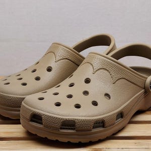 May include: A pair of tan-colored Crocs clogs. The shoes have multiple ventilation holes and a textured design around the edges. The clogs are made of a rubbery material and are sitting on a wooden surface.
