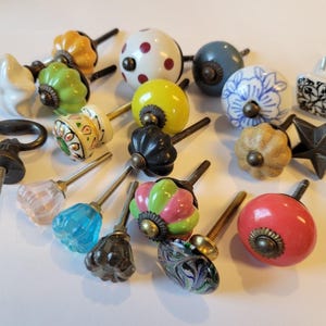 May include: A selection of decorative cabinet knobs in diverse colours and designs. The knobs feature various shapes, including round, star, and floral, and are made from materials like ceramic and metal. Colours include red, blue, green, yellow, and white.