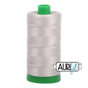 May include: A cylindrical spool of light gray sewing thread featuring a bright green plastic top and base. The spool is wrapped in smooth, tightly wound thread and displays the brand name AURIfil in black and blue text on the lower right side.