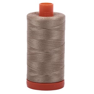 May include: A spool of taupe sewing thread featuring a cylindrical shape with a smooth, tightly wound surface. The spool has a bright orange plastic top and a matching orange plastic base with the embossed text AURIFIL visible along the edge.