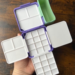 Puede incluir: Una paleta de acuarela morada y blanca con múltiples compartimentos y un recipiente verde. La paleta está abierta, revelando varias secciones para mezclar y contener pintura. La paleta está diseñada para artistas.