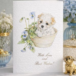 Puede incluir: Tarjeta de felicitación con una ilustración a la acuarela de un cachorro blanco y esponjoso en una maceta beige con flores azules. La tarjeta incluye el texto "With Love and Best Wishes!" en escritura elegante.