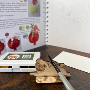 May include: A wooden pug-shaped coaster with a floral design, a watercolor paint set, a paintbrush, and an open spiral-bound book with pomegranate illustrations and instructions. The book also has a blank page with a pomegranate outline.