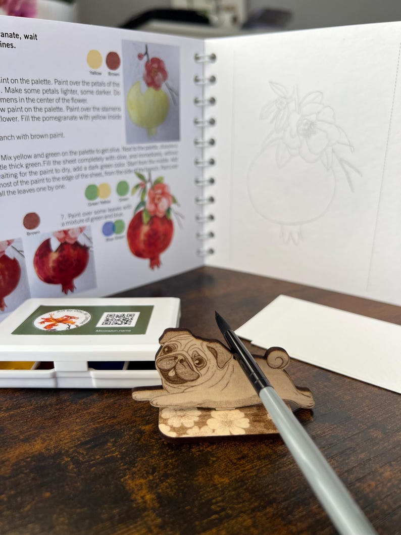 May include: A wooden pug-shaped coaster with a floral design, a watercolor paint set, a paintbrush, and an open spiral-bound book with pomegranate illustrations and instructions. The book also has a blank page with a pomegranate outline.