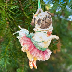 May include: A wooden pug dog ornament in a ballerina outfit. The pug is wearing a pink tutu, green sash, and ballet slippers. It has a floral crown and is hanging from a green and white ribbon. The ornament is set against a backdrop of green pine needles.