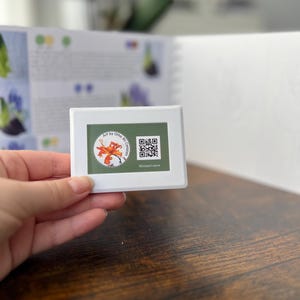 May include: A white rectangular object with a floral design and the text "Art by Olha Mi Coro" and a QR code. The object is held in a hand, with a book and wooden surface in the background.