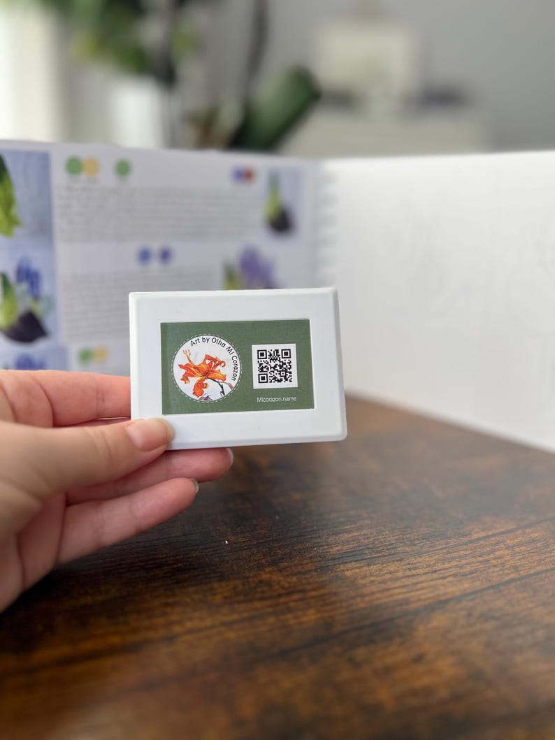 May include: A white rectangular object with a floral design and the text "Art by Olha Mi Coro" and a QR code. The object is held in a hand, with a book and wooden surface in the background.