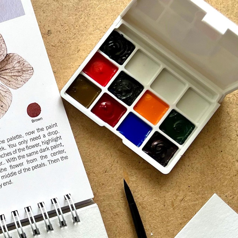 May include: A white watercolor paint palette with a variety of colors, including black, red, orange, and blue. A small paintbrush and a notepad with text are also visible. The palette is open, revealing the vibrant colors.