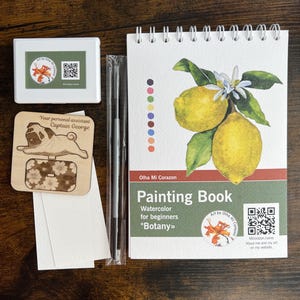 May include: A watercolor painting kit featuring a spiral-bound painting book with a lemon illustration, a wooden pug-shaped tag, a white box with a floral logo, a paintbrush, and watercolor paper. The book is titled "Painting Book" and includes the text "Botany".