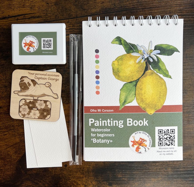 May include: A watercolor painting kit featuring a spiral-bound painting book with a lemon illustration, a wooden pug-shaped tag, a white box with a floral logo, a paintbrush, and watercolor paper. The book is titled "Painting Book" and includes the text "Botany".