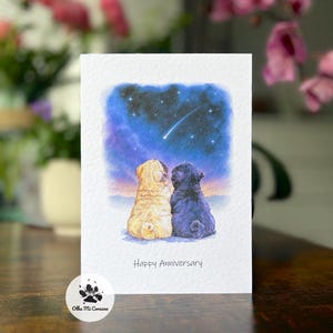 May include: A greetings card with a watercolour illustration of two pugs, one tan and one blue, looking at a starry night sky with a shooting star. The words "Happy Anniversary" are printed below the image. A small logo is in the lower left corner.
