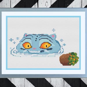 May include: Cross-stitch art of a blue cat with orange eyes and a wide grin, emerging from water. A brown pot with green and orange foliage sits to the right. The artwork is framed in a light blue and silver frame.