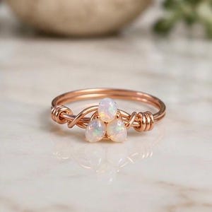 May include: A rose gold wire-wrapped ring featuring three small, iridescent, oval-shaped opals arranged in a triangular cluster at the center. The band is constructed from thin, coiled metal wire that wraps around the base of the stones.