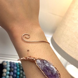 May include: A copper wire cuff bracelet featuring a raw, elongated purple amethyst crystal wrapped in coiled copper wire. The bracelet has a simple spiral design at one end and a secure wire-wrapped setting for the stone, creating a metallic look.