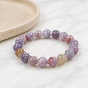 Könnte beinhalten: Ein elastisches Armband aus glatten, runden Perlen in verschiedenen Schattierungen von Violett, Lavendel und sanftem Goldgelb. Die Perlen haben eine polierte, halbtransparente Oberfläche mit sichtbaren natürlichen mineralischen Einschlüssen.