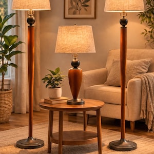 Puede incluir: Tres lámparas con pantallas de tela color beige y bases de madera oscura iluminan una sala de estar. Dos son lámparas de pie y una es una lámpara de mesa sobre una mesa auxiliar redonda. Una impresión botánica enmarcada cuelga de la pared.
