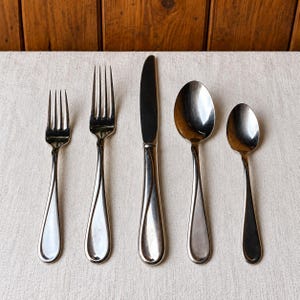 May include: A five-piece stainless steel flatware set consisting of a small fork, a large fork, a dinner knife, a large spoon, and a small spoon. Each piece features a smooth, polished finish with a subtle curved design on the handle.