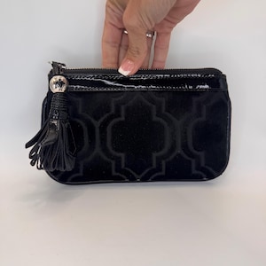 May include: Black clutch bag with a velvet geometric pattern. The bag has a patent leather zip and a decorative tassel. The bag is being held by a person.