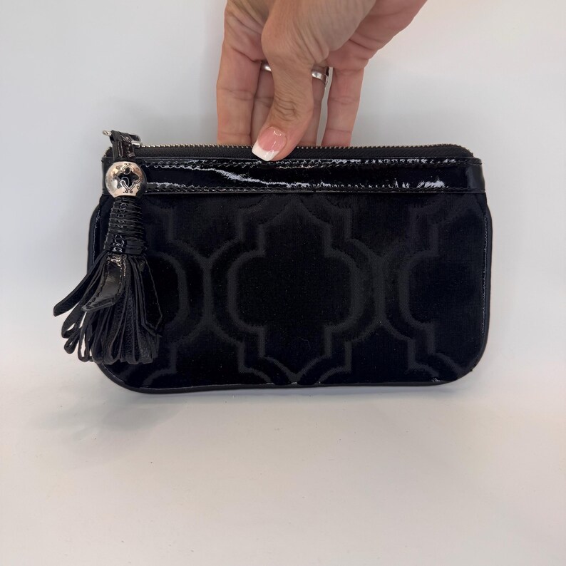 May include: Black clutch bag with a velvet geometric pattern. The bag has a patent leather zip and a decorative tassel. The bag is being held by a person.