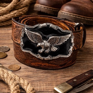 Può includere: Cintura in pelle marrone con fibbia argentata raffigurante un'aquila su sfondo nero. Posizionata su una superficie di legno, insieme a stivali marroni, monete e una corda, crea un'atmosfera rustica.