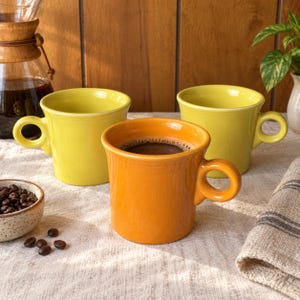 Op de afbeelding: Drie keramische mokken met ronde oren, waaronder een oranje mok gevuld met zwarte koffie en twee gele mokken. De mokken hebben een gladde, glanzende afwerking en een eenvoudige, cilindrische vorm. Een klein schaaltje met koffiebonen staat ernaast.