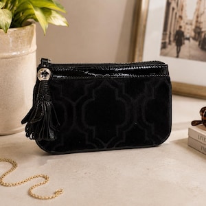 May include: Black velvet clutch bag with a geometric pattern and a patent leather top. The bag features a tassel and a silver-tone zipper pull. A gold chain, sunglasses, and books are also in the image.