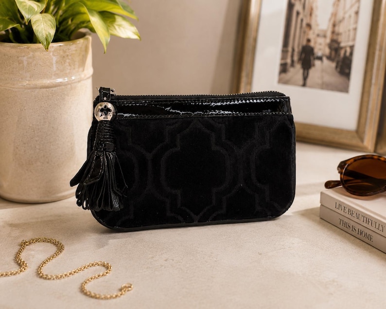 May include: Black velvet clutch bag with a geometric pattern and a patent leather top. The bag features a tassel and a silver-tone zipper pull. A gold chain, sunglasses, and books are also in the image.